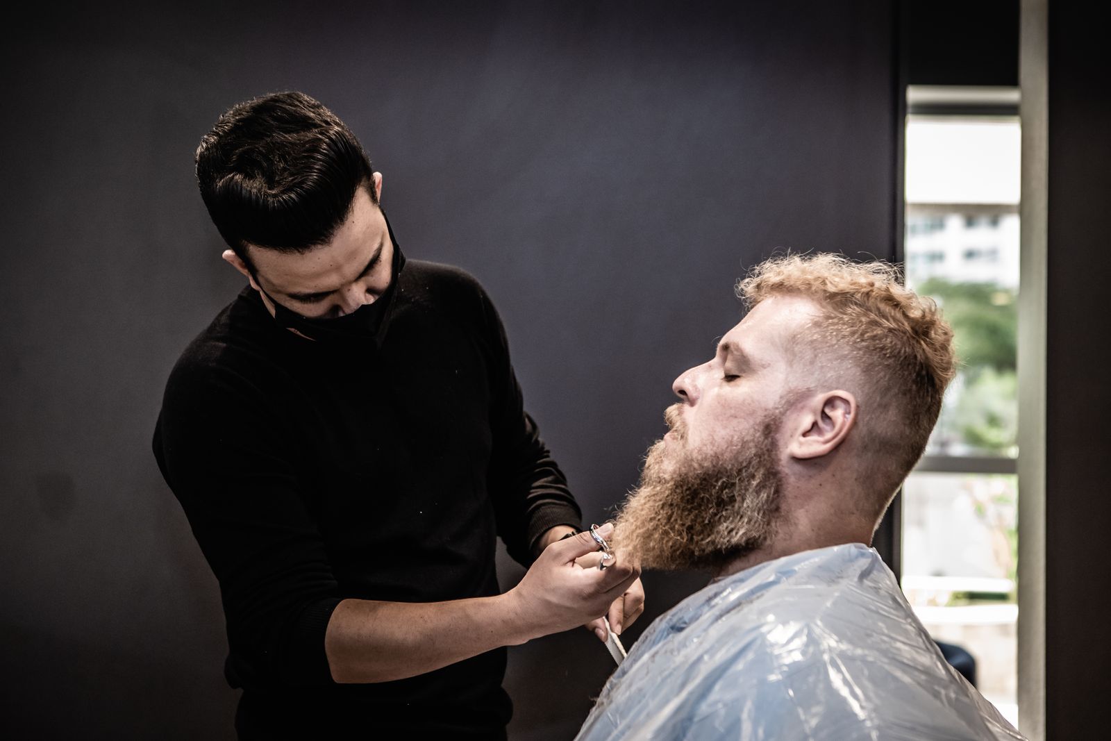 Barber shaping a full beard with scissors in a dark shop