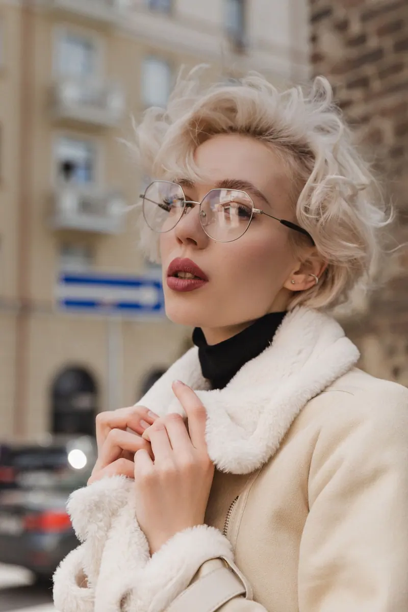 Portrait of a fashionable woman with blond hair and eyeglasses wearing a fluffy jacket outdoors.