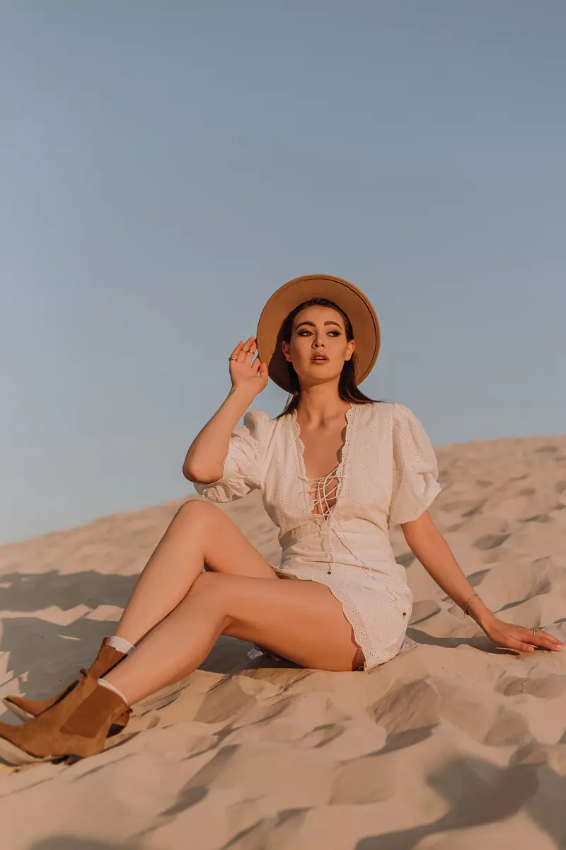 Stylish woman wearing a hat and dress sitting on sand dunes at sunset.