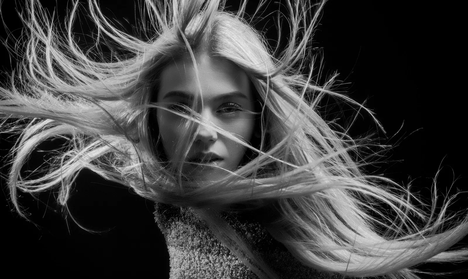 Black-and-white portrait of a woman with long platinum hair flying across her face against a black backdrop