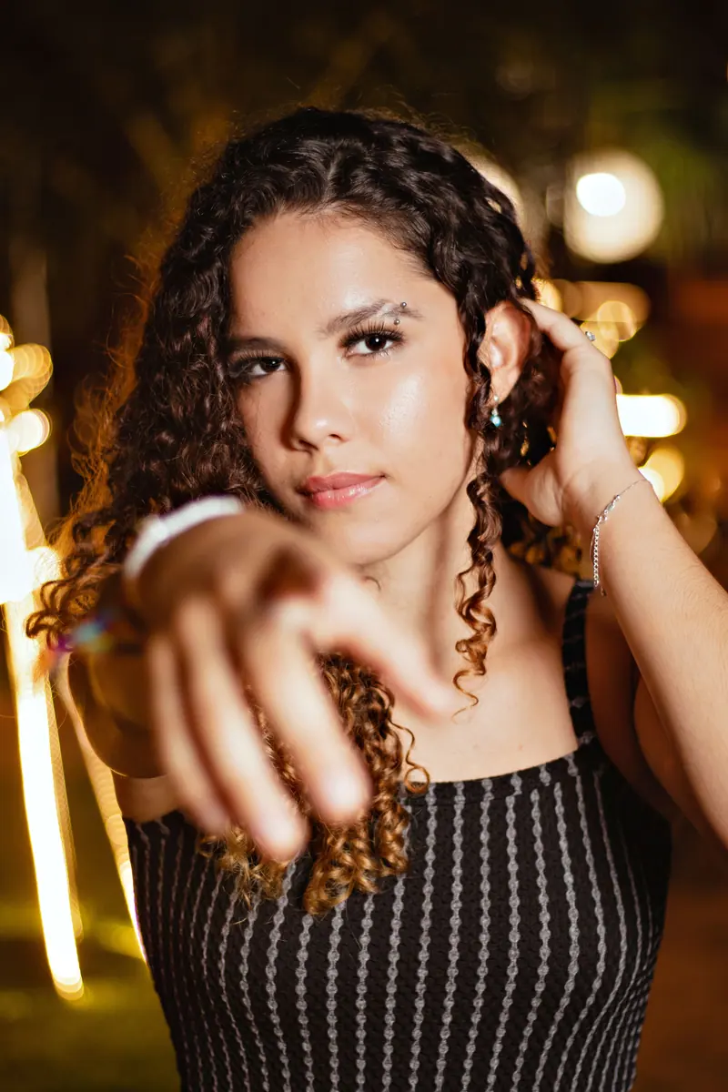 A young woman with curly hair pointing at the camera in a blurred background setting.