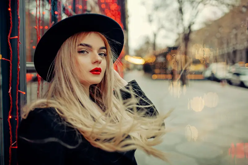 Elegant woman with bold red lips and black hat posing outdoors, capturing a chic urban vibe.