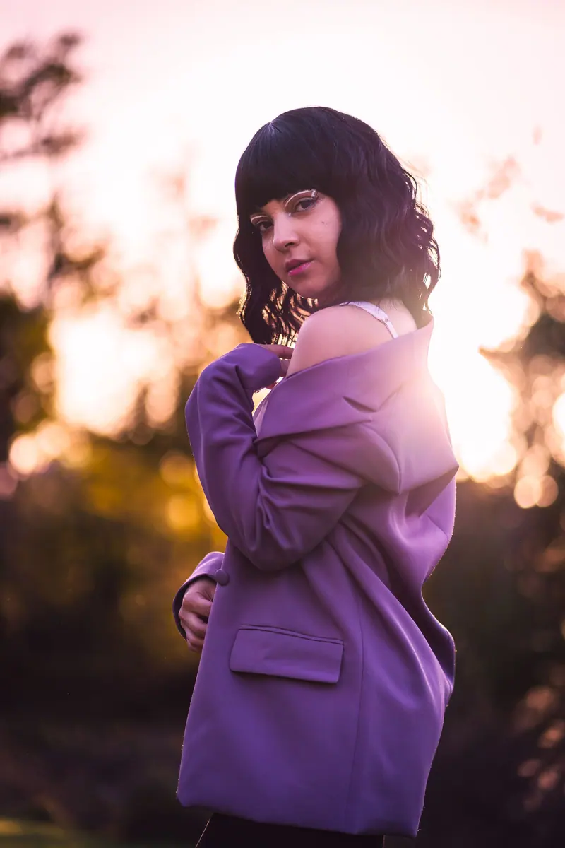 Elegant woman in a purple coat posing outdoors during sunset with a stylish flair.