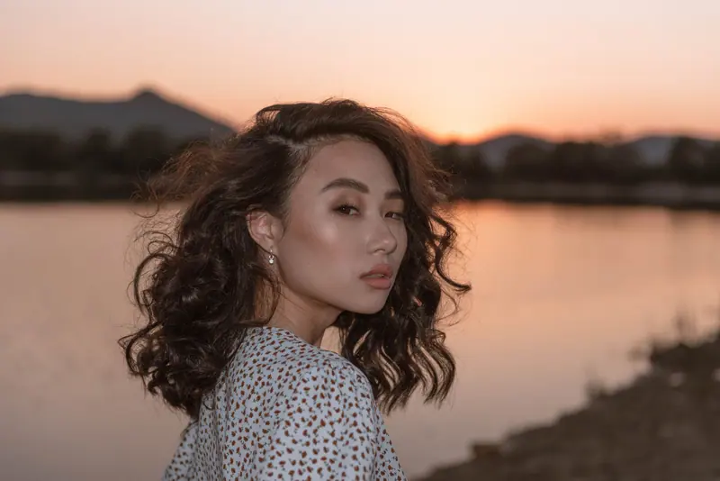 A woman with curly hair in a floral top looks over her shoulder at sunset by the lake.