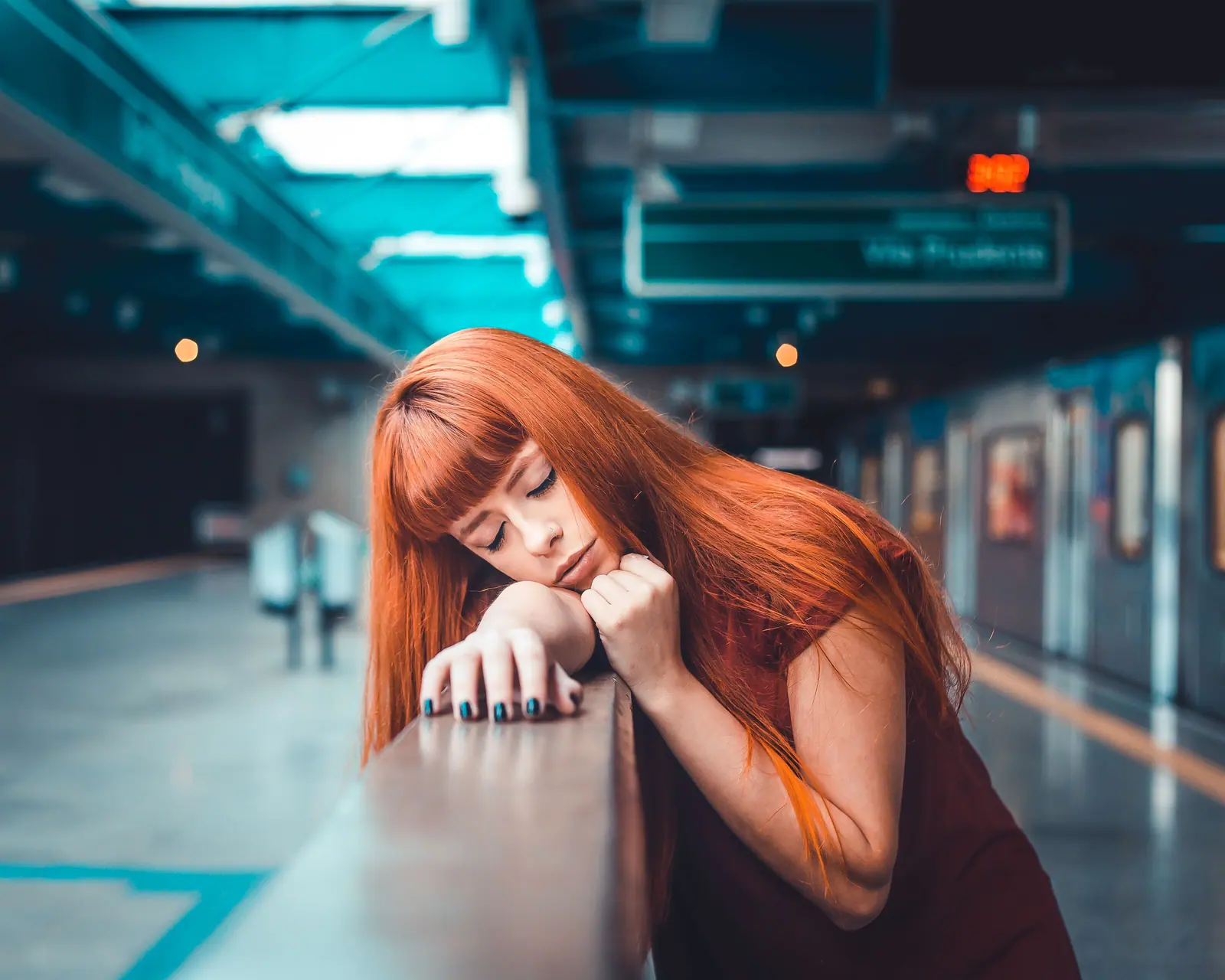 Woman with long copper hair resting her chin on a railing in a blue-lit station at night