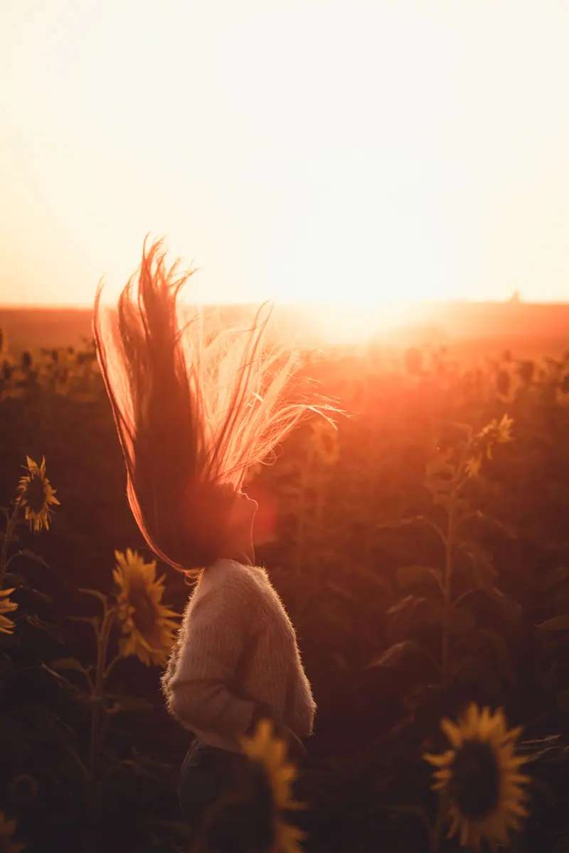 A woman flips her hair in a sunflower field during sunset, creating a silhouette effect.