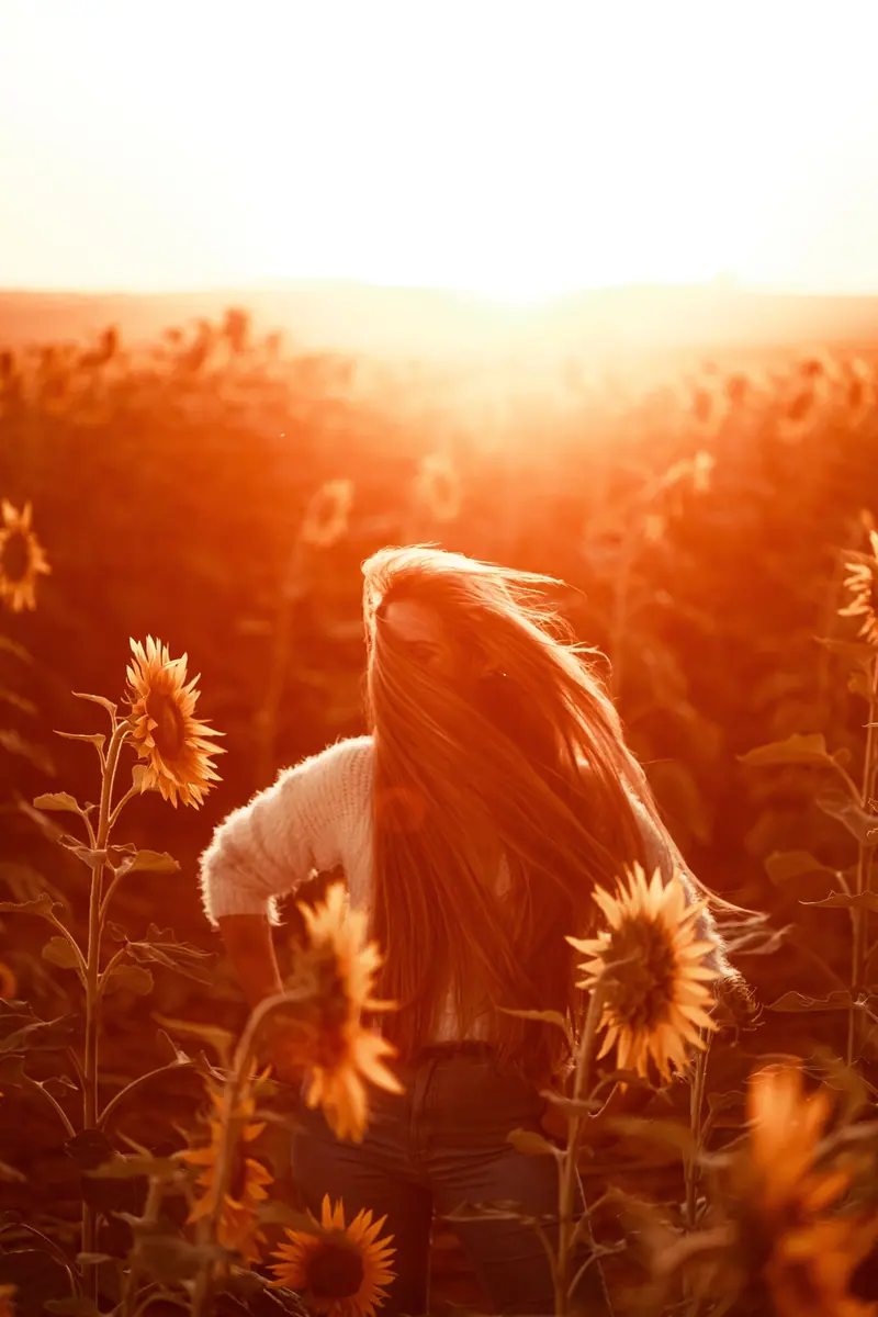 A woman with long hair stands in a sunflower field at sunset, bathed in golden light.