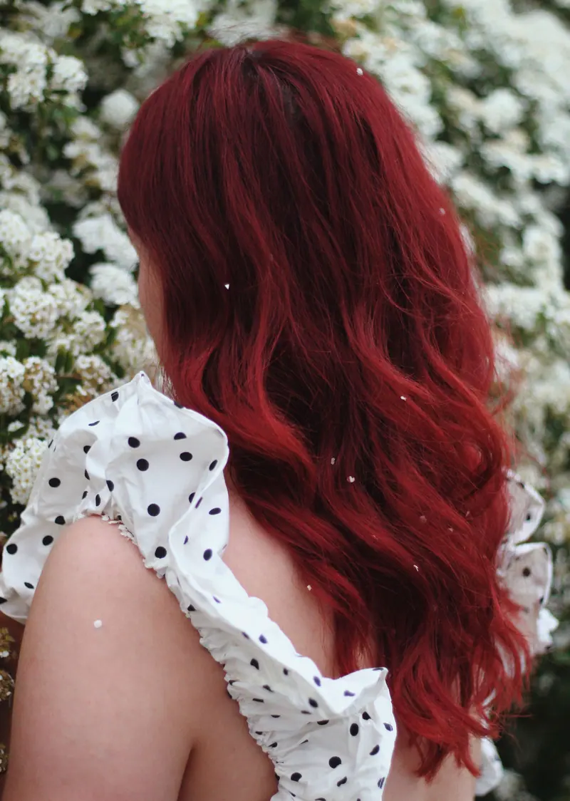 Back view of a woman with long red hair in a polka dot dress standing near white flowers.