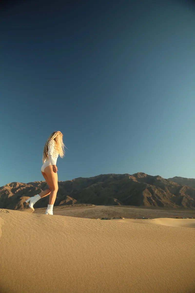 Blonde woman walking in a vast desert landscape with mountains under a clear sky.