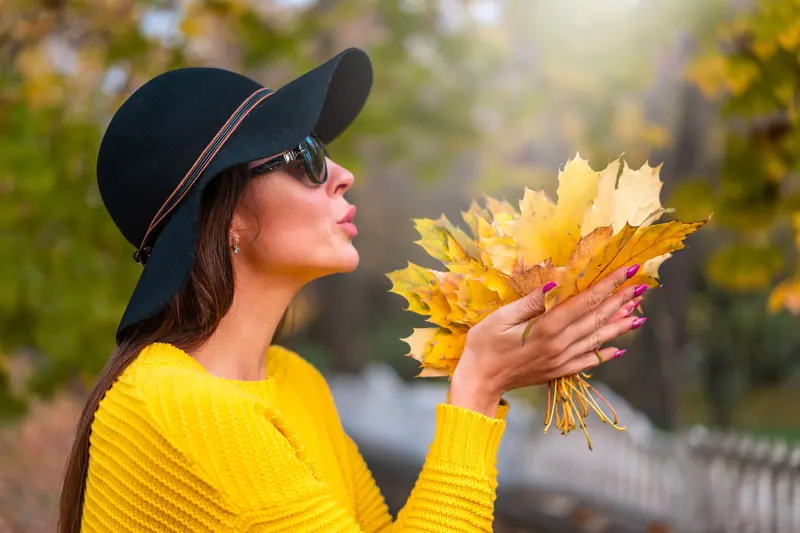 Woman in yellow sweater and hat holding autumn leaves outdoors.