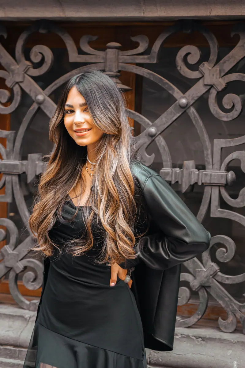 Confident woman in a black dress and leather jacket posing stylishly against a decorative metal backdrop.