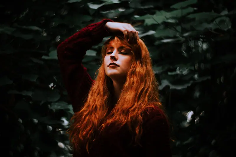 Artistic portrait of a woman with auburn hair, eyes closed, in a natural outdoor setting.