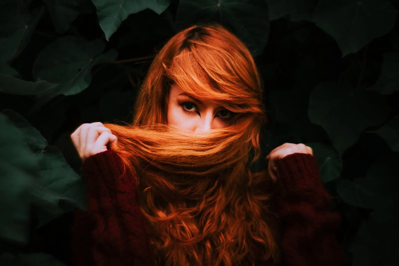 A captivating portrait of a woman with auburn hair partially covering her face, surrounded by lush green leaves.