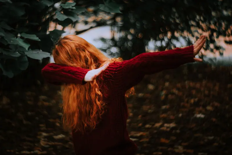 A redheaded woman wearing a red sweater dabs amidst a forest setting, with autumn leaves.