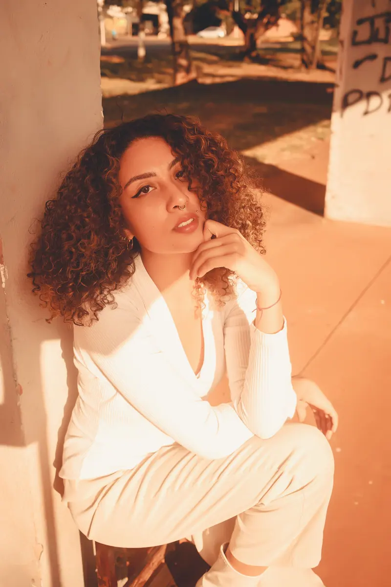 A fashionable woman with curly hair poses outdoors in warm sunlight, dressed in white.