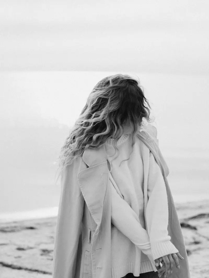 A woman with wavy hair stands on a peaceful beach, captured in a stylish black and white photo.
