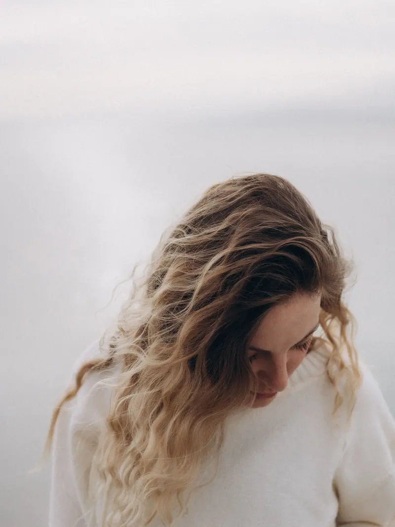 Young woman with blonde curly hair in white sweater looking down in a serene outdoor setting.