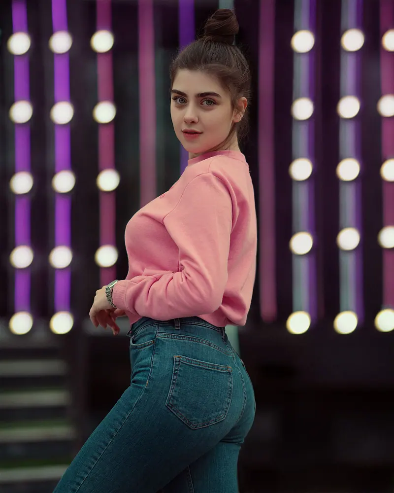 Fashionable young woman in pink sweater posing with lights in the background.