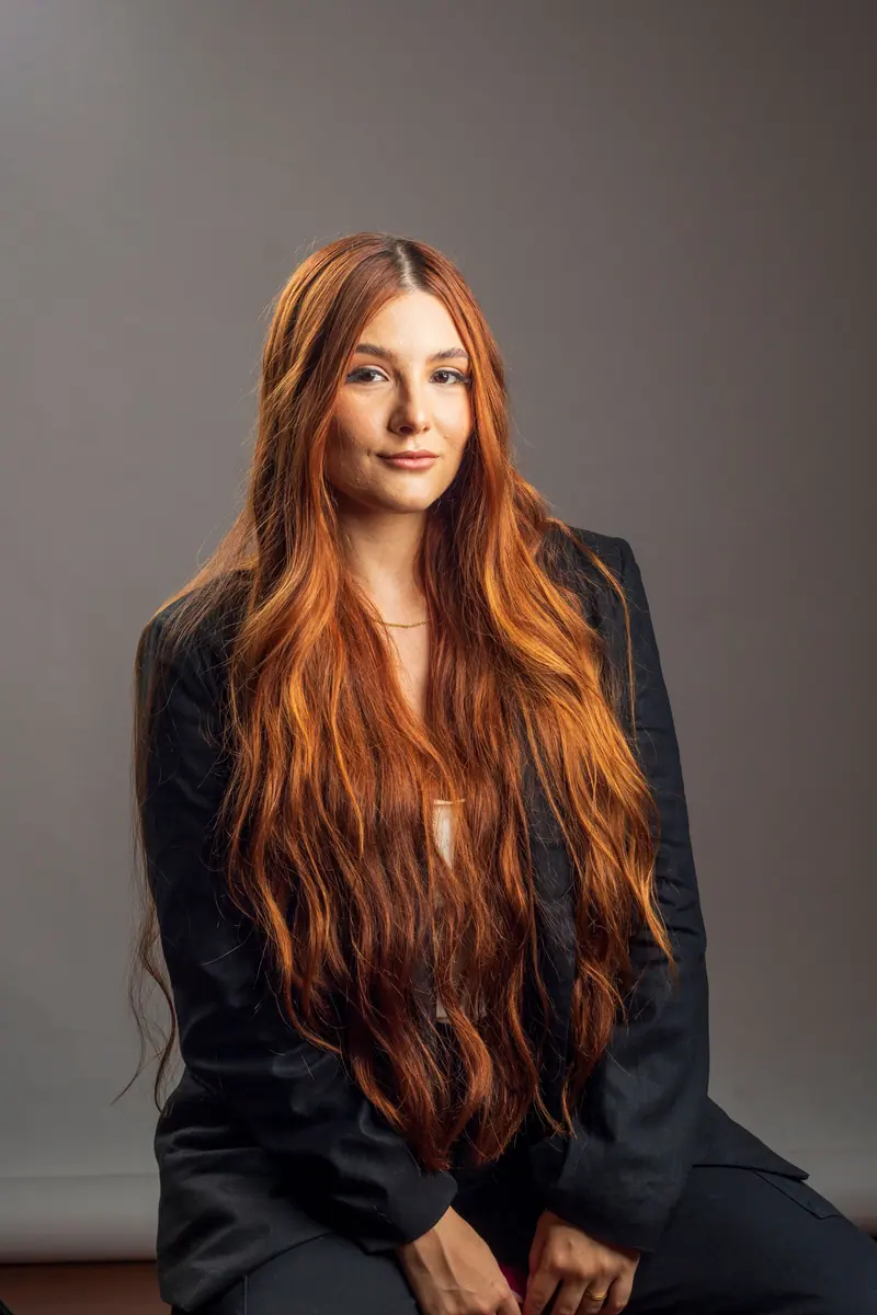 A beautiful portrait of a woman with long red hair sitting gracefully against a neutral backdrop.