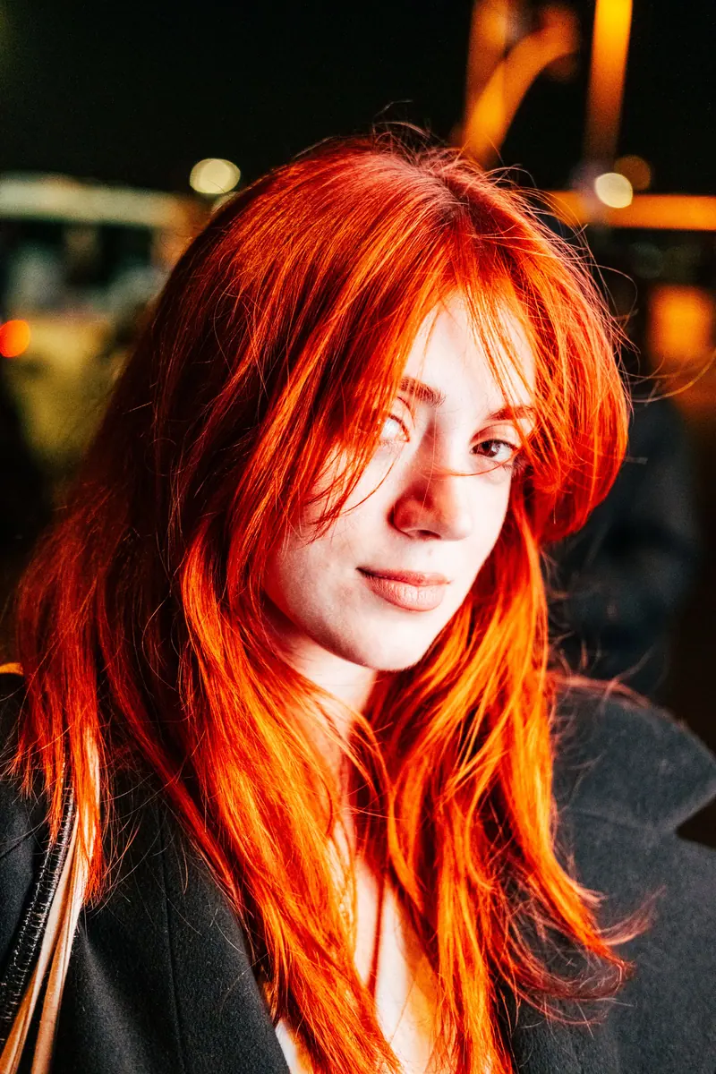 Portrait of a woman with striking red hair and a confident smile, captured at night outdoors.