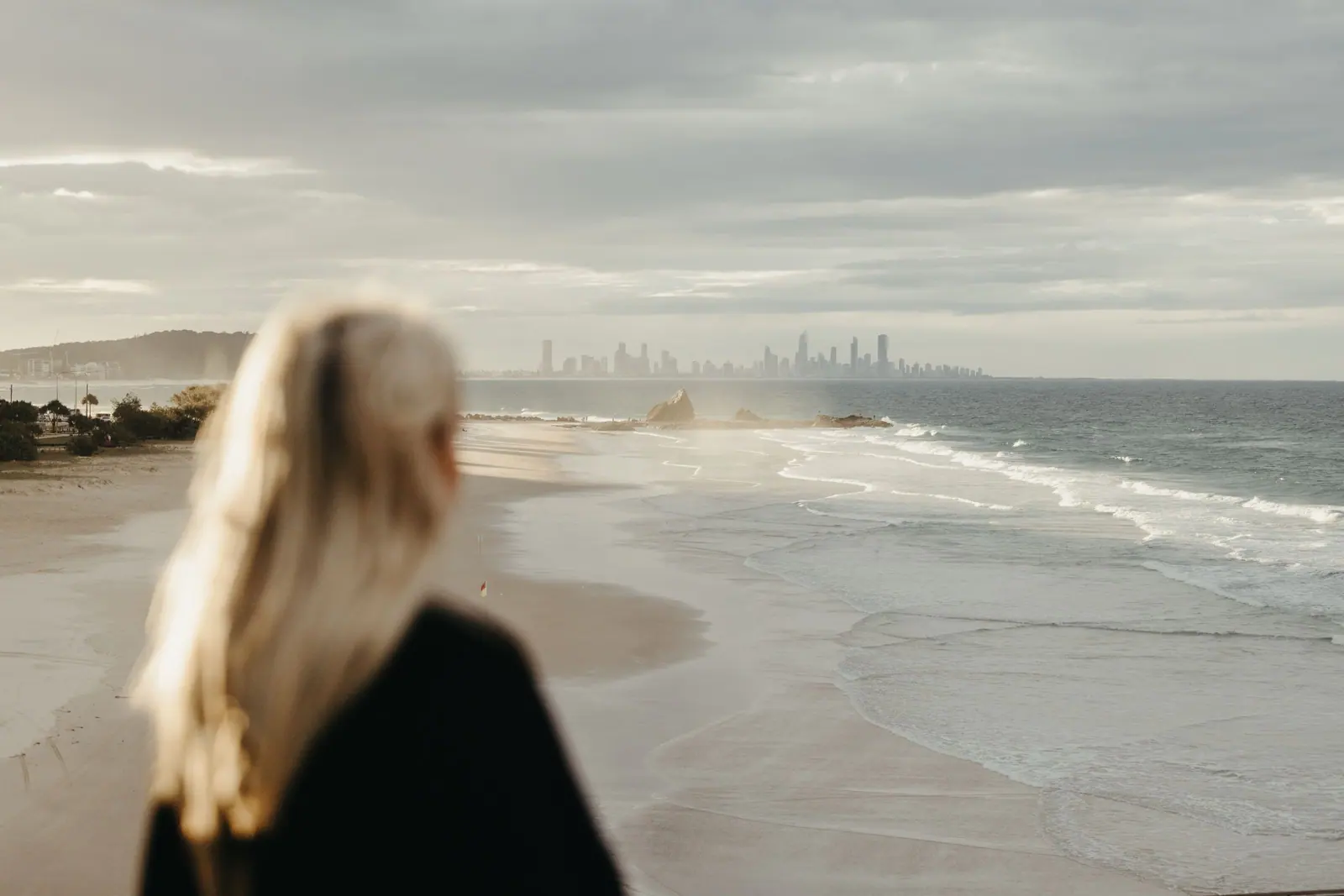 Blonde woman stands on Currumbin Beach, Gold Coast with city skyline in view.