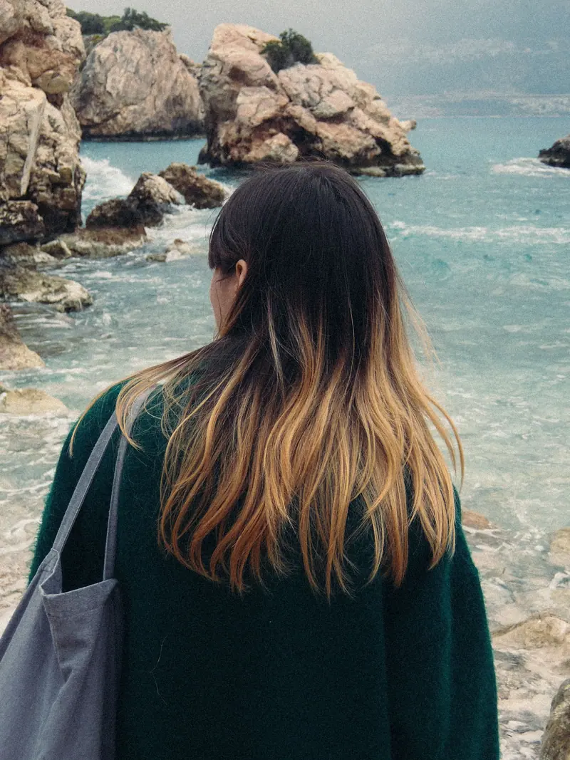 A woman with ombre hair stands by the rocky seaside, embracing tranquility and leisure.