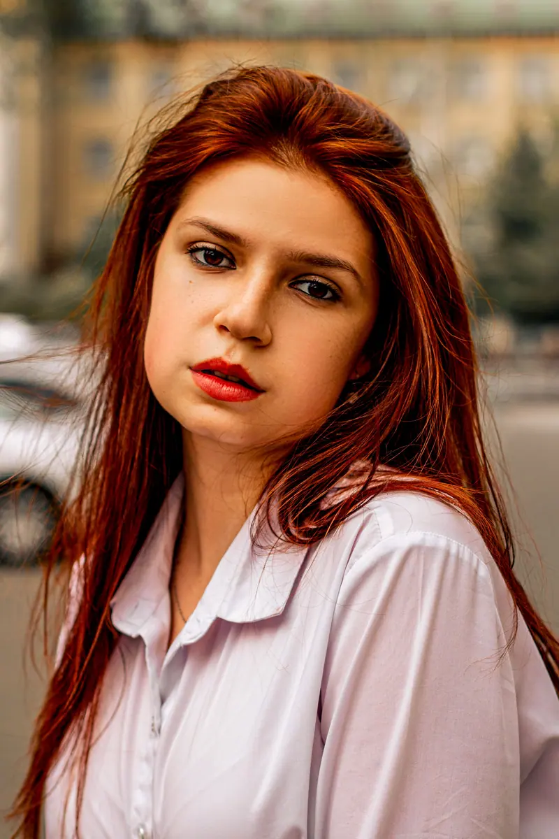 Striking outdoor portrait of a young, red-haired woman in a white shirt with a serious expression.