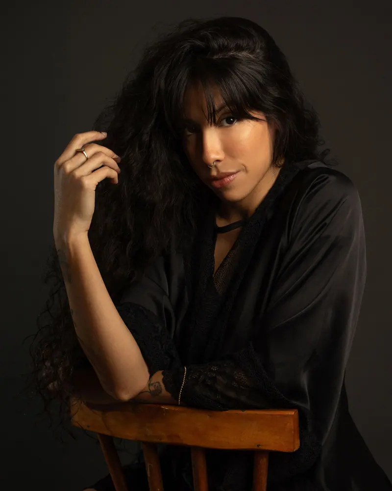 Portrait of a woman in black nightwear, with long wavy hair, sitting indoors.