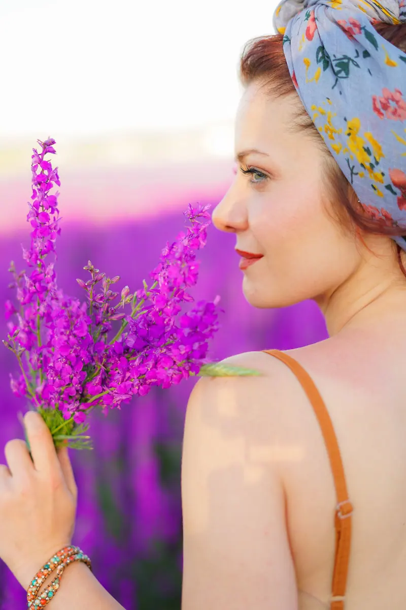 A woman with red hair and floral scarf holding lavender in a blooming lavender field during summer.