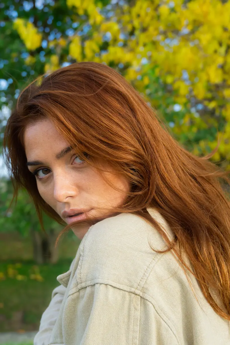 A young woman with auburn hair looks over her shoulder in a lush park setting.