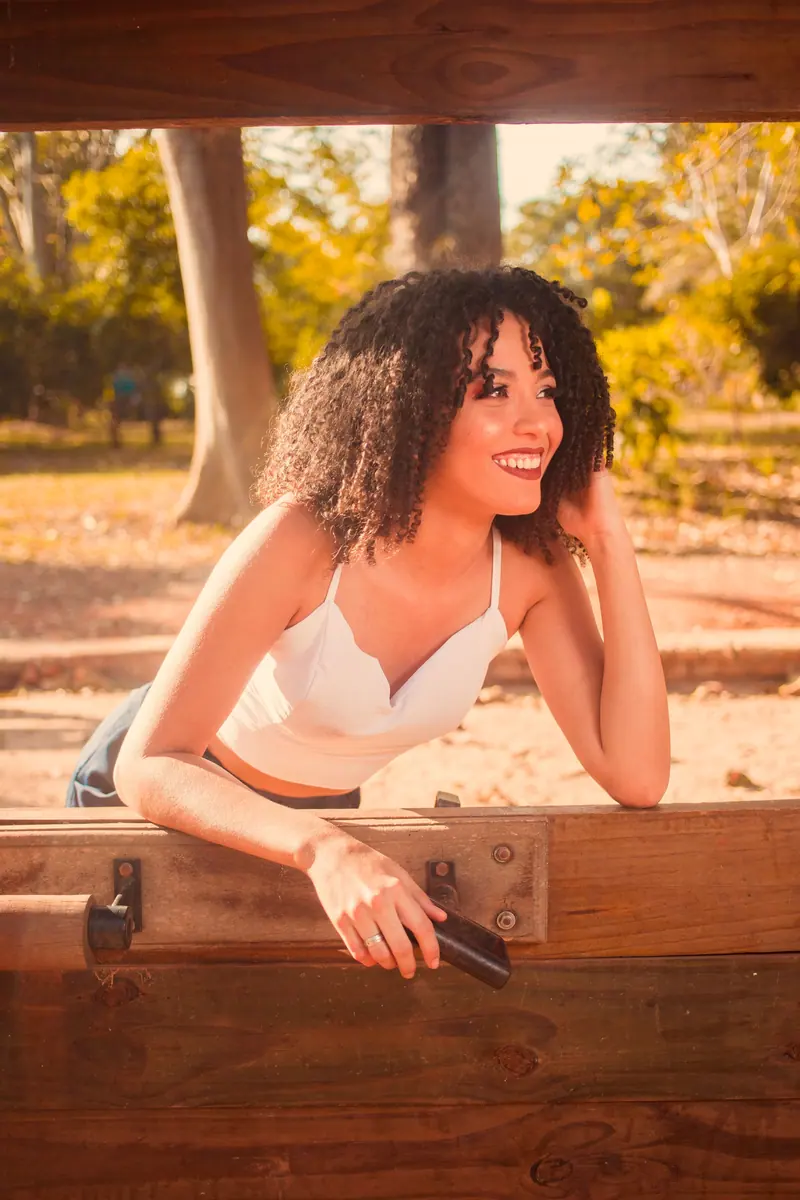 A cheerful young woman with curly hair enjoys a sunny day outdoors in a rural setting.
