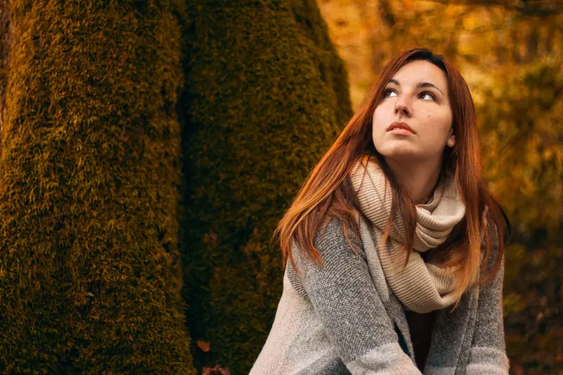 Red-haired woman gazing upward with autumn foliage in background. Cozy and contemplative.