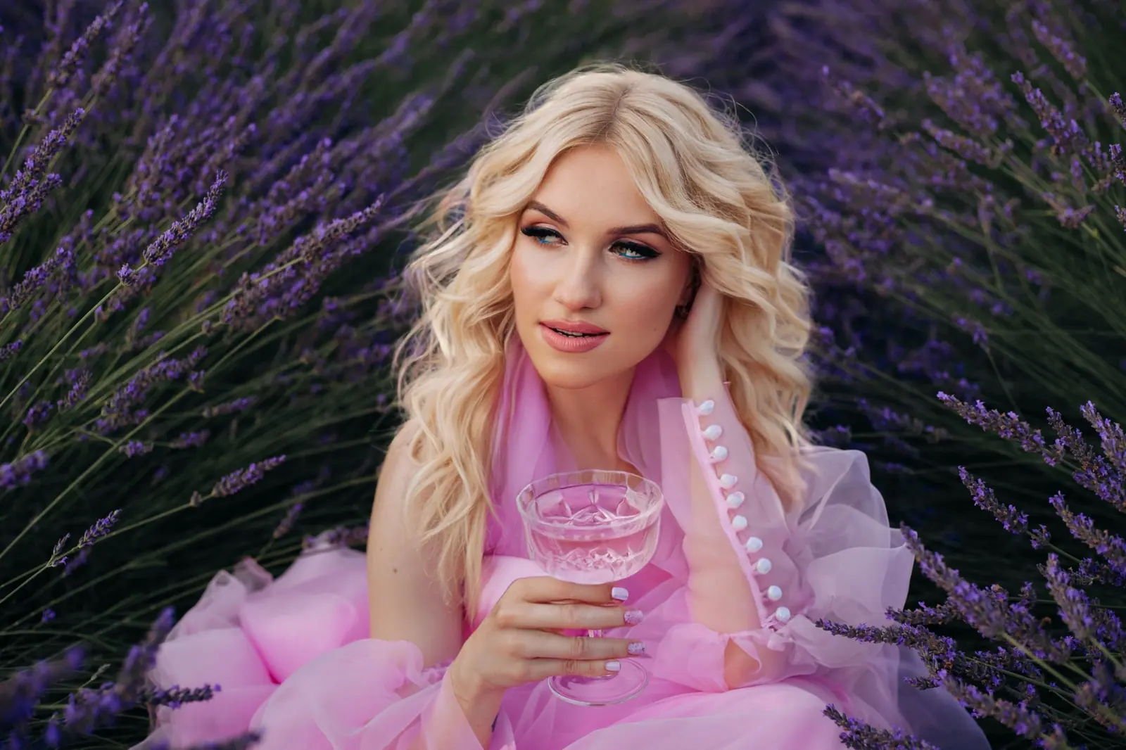 Blonde woman with soft waves sitting in a field of lavender holding a drink, dressed in lilac tulle