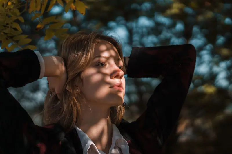 A young Caucasian woman posing outdoors under dappled sunlight in a serene, natural setting.