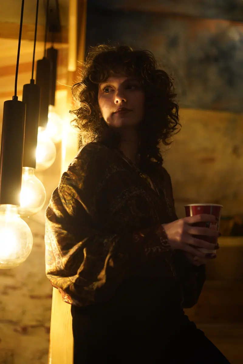 Woman with curly hair holding a cup, illuminated by warm hanging bulbs in a cozy setting.