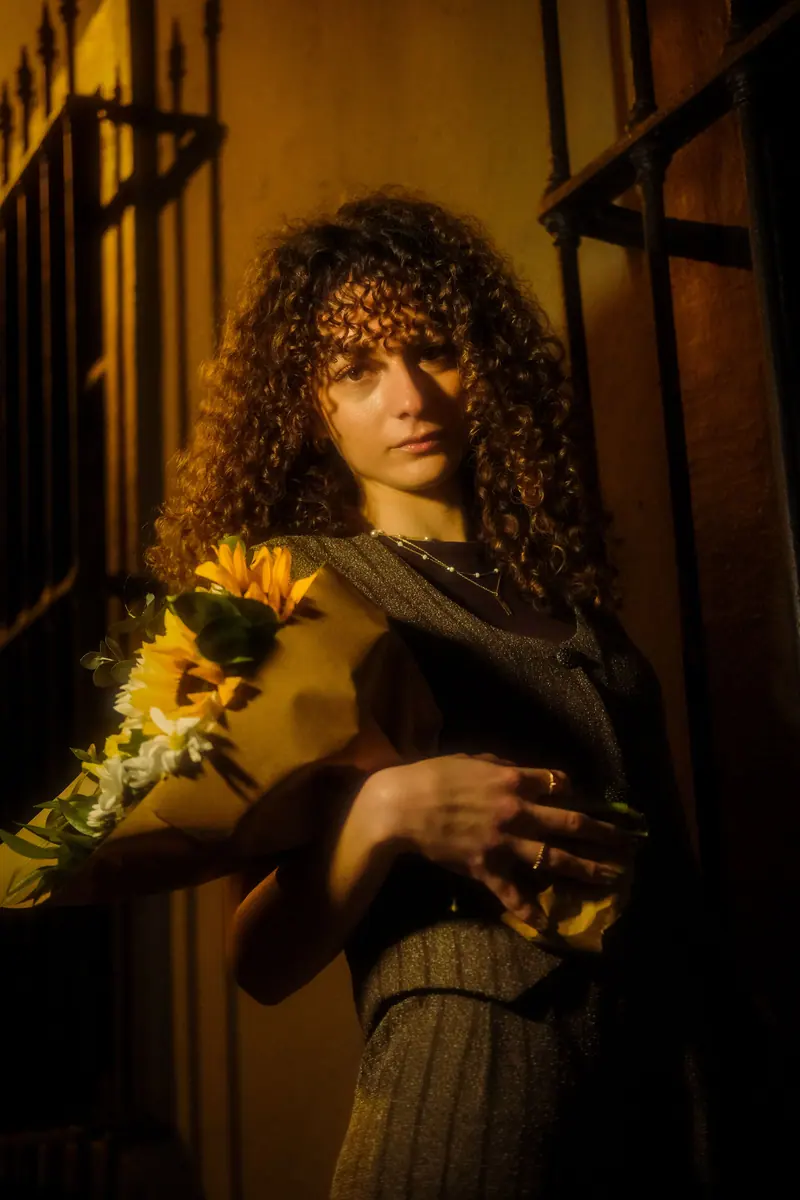 A woman with curly hair holds a sunflower bouquet against a dimly lit urban wall.