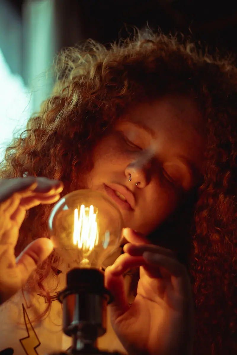 Warm portrait of a woman with curly hair holding an illuminated retro light bulb indoors.