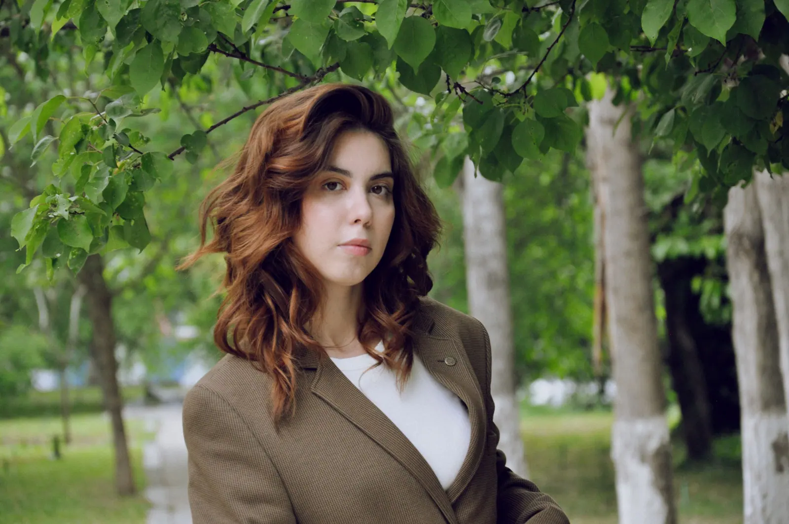 Woman with long brunette balayage waves in a tailored brown blazer standing among birch trees