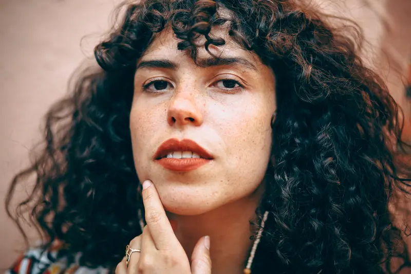 Close-up portrait of a woman with curly hair, freckles, and thoughtful expression.