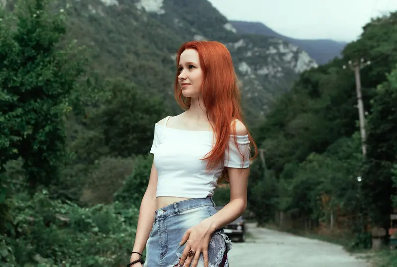 A young woman with red hair stands outdoors in a scenic mountain landscape, embracing summer fashion.