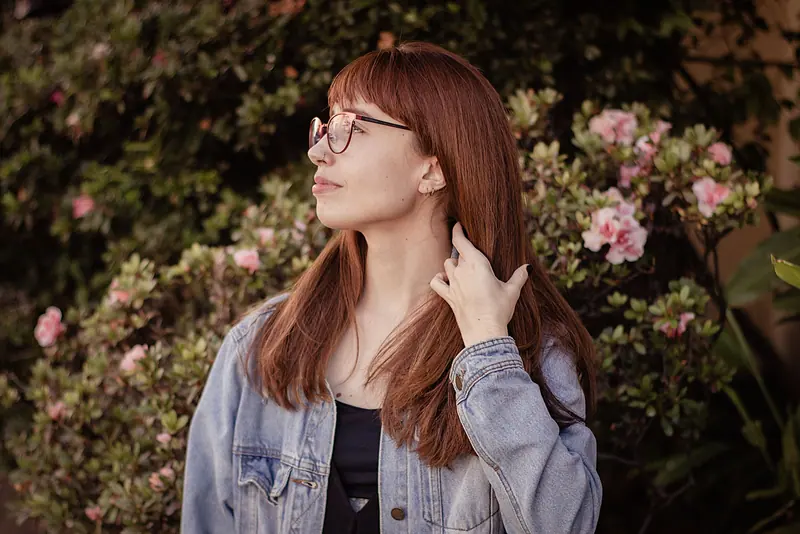 Redhead woman in denim jacket poses outdoors against pink floral backdrop.