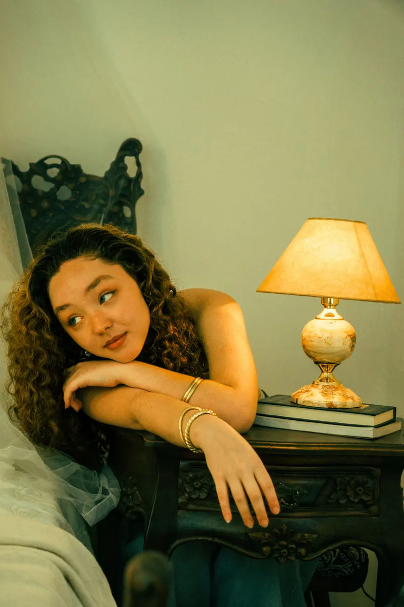 Young woman leaning on a bedside table with a lamp and books, capturing a cozy indoor scene.
