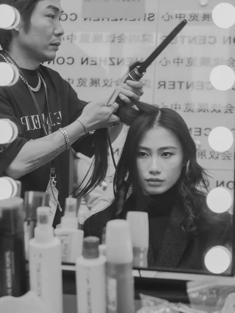 A stylist curling hair in a backstage dressing room with a mirror setup.