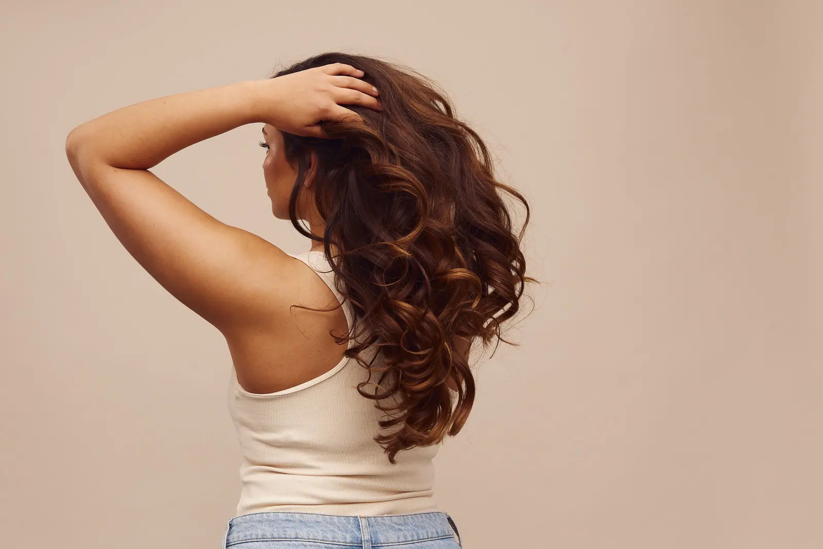 Back view of a woman running a hand through long, glossy chestnut curls against a warm oat backdrop
