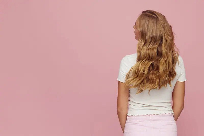 Rear view of woman with long blonde wavy hair against pink backdrop.