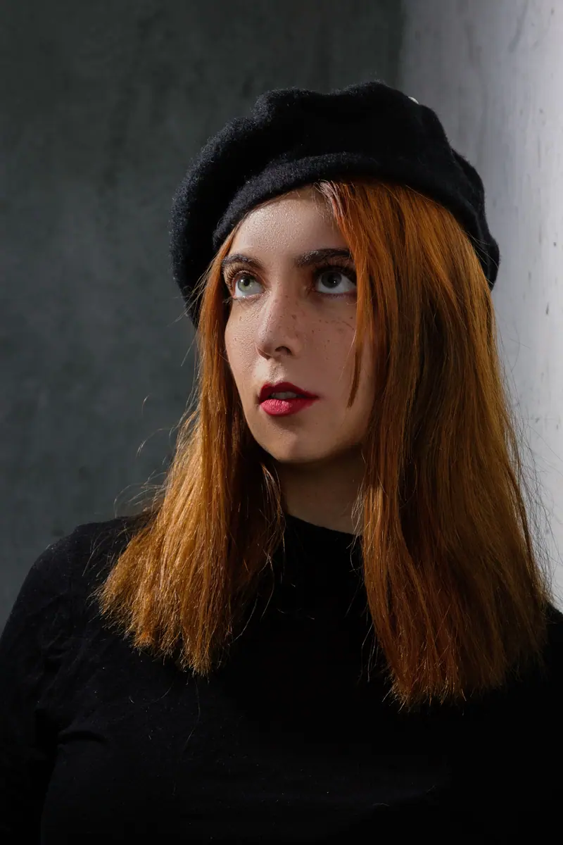 Stylish portrait of a redhead woman with freckles in a black beret and top against a gray background.