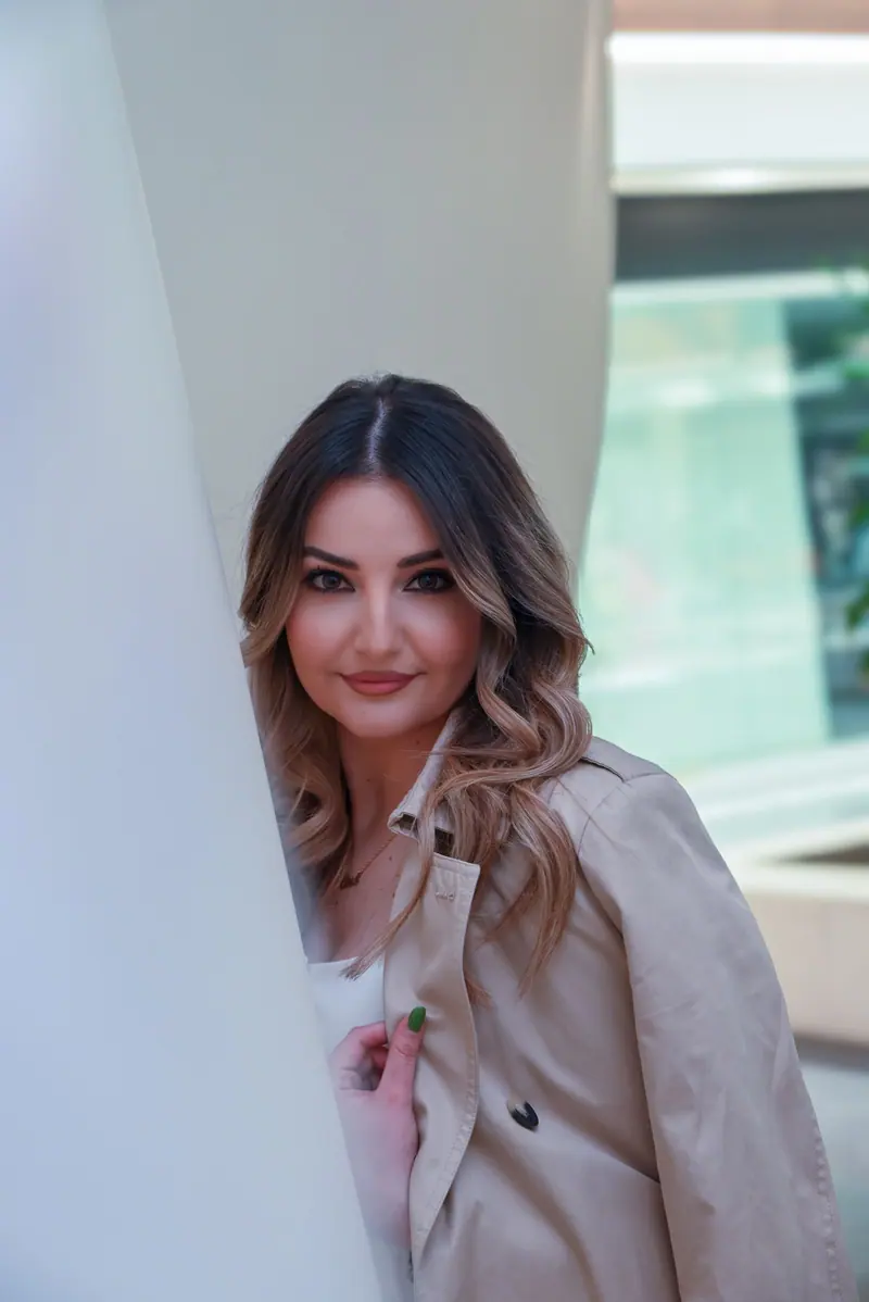 Stylish woman wearing a beige trench coat posing outdoors with a confident smile.
