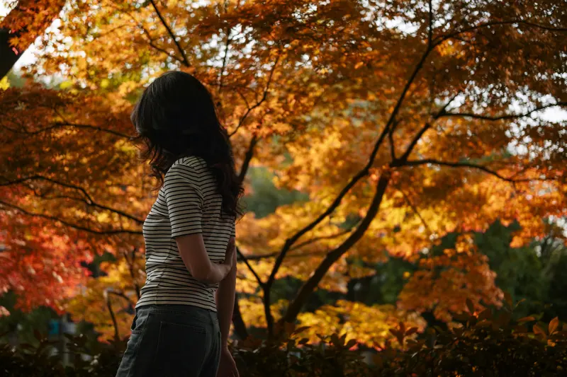 A woman in a striped shirt stands amidst vibrant autumn leaves, creating a serene and picturesque scene.