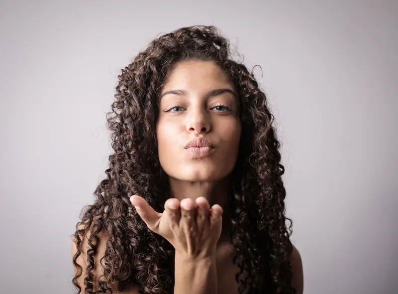 Elegant young woman with curly hair sending a playful kiss in studio. Perfect for beauty and lifestyle themes.