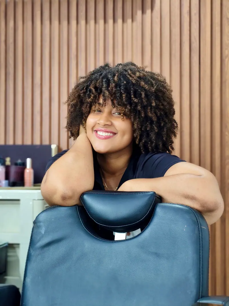 Woman with curly hair smiles while leaning on a salon chair, creating a cheerful and welcoming atmosphere.